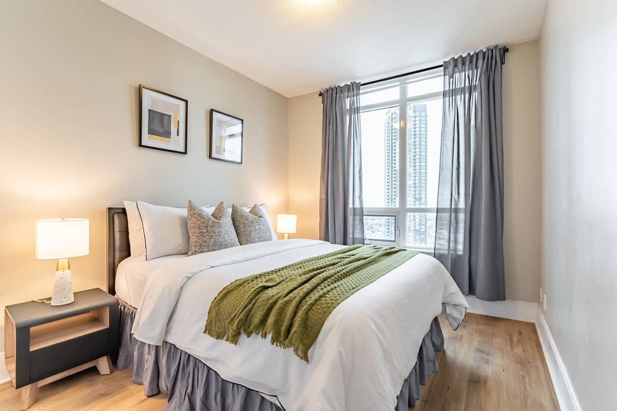 A bedroom with a bed, two bedside tables, a window with curtains, and framed pictures on the wall.