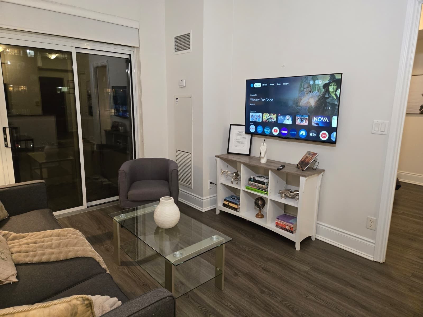 A modern living room with a glass coffee table, a grey armchair, and a flat-screen TV mounted on the wall displaying a movie.