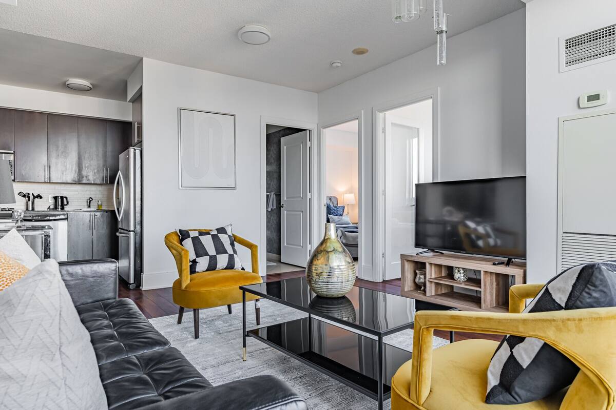 A modern living room with a sofa, yellow armchair, and a TV. There is a kitchen area with cabinets and a refrigerator.