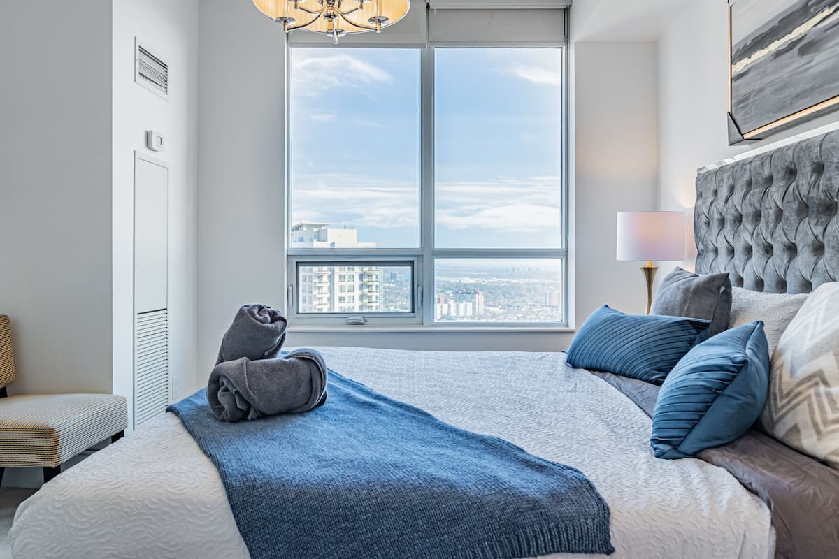 A modern hotel room with a large bed, a view of the city, a tufted headboard, and a bedside lamp.
