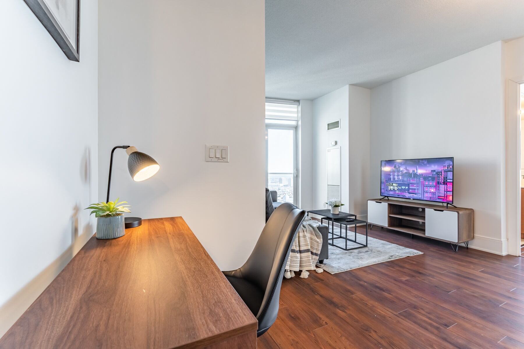 A modern living room with a wooden dining table, a black chair, a TV on a wooden stand, and a potted plant.