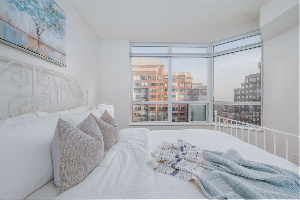 A bedroom with a large window, a bed with white bedding, and a painting on the wall.
