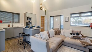 Een moderne keuken met een centraal eiland, een zithoek met een bank, en uitzicht op buiten door grote ramen.