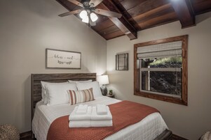 A neatly made bed with white bedding and a red blanket, a ceiling fan, a framed inspirational quote on the wall, a window with a view of greenery, and a small table with a lamp and a plant.