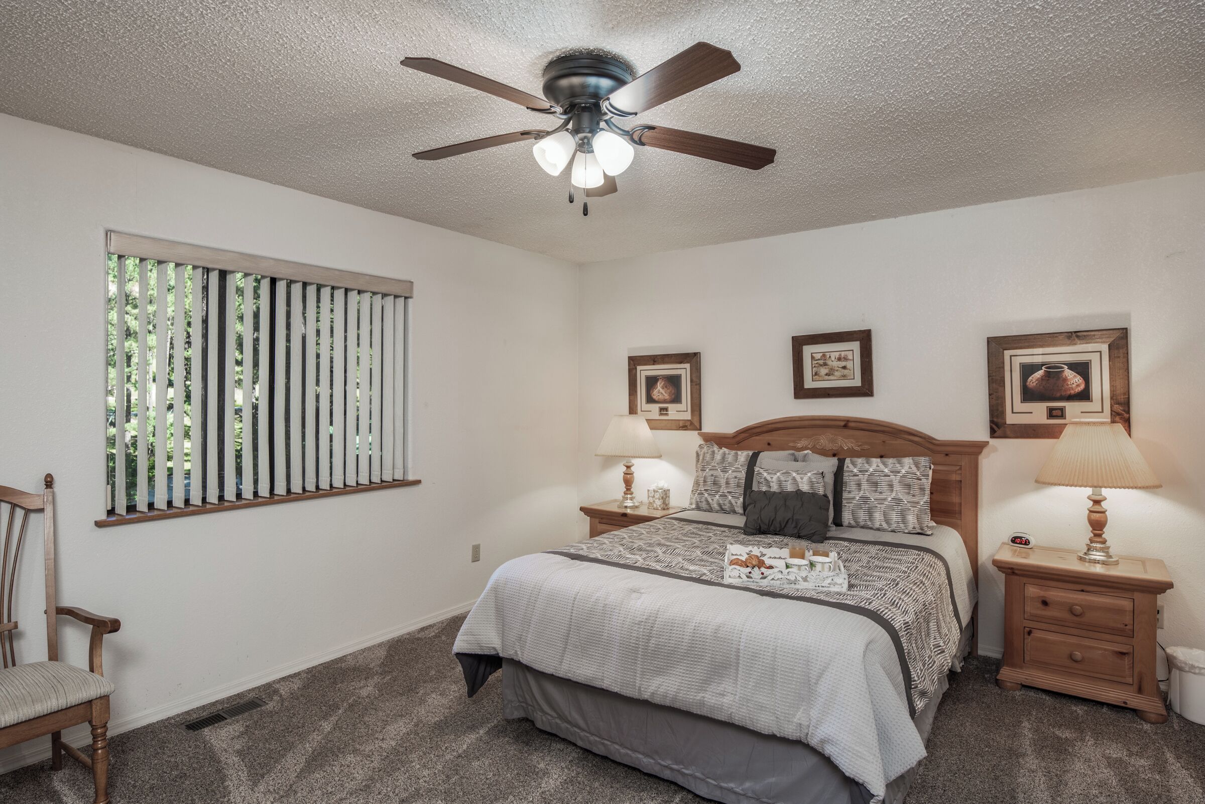 A bedroom with a bed, two bedside tables, a ceiling fan, a window with blinds, and framed pictures on the wall.