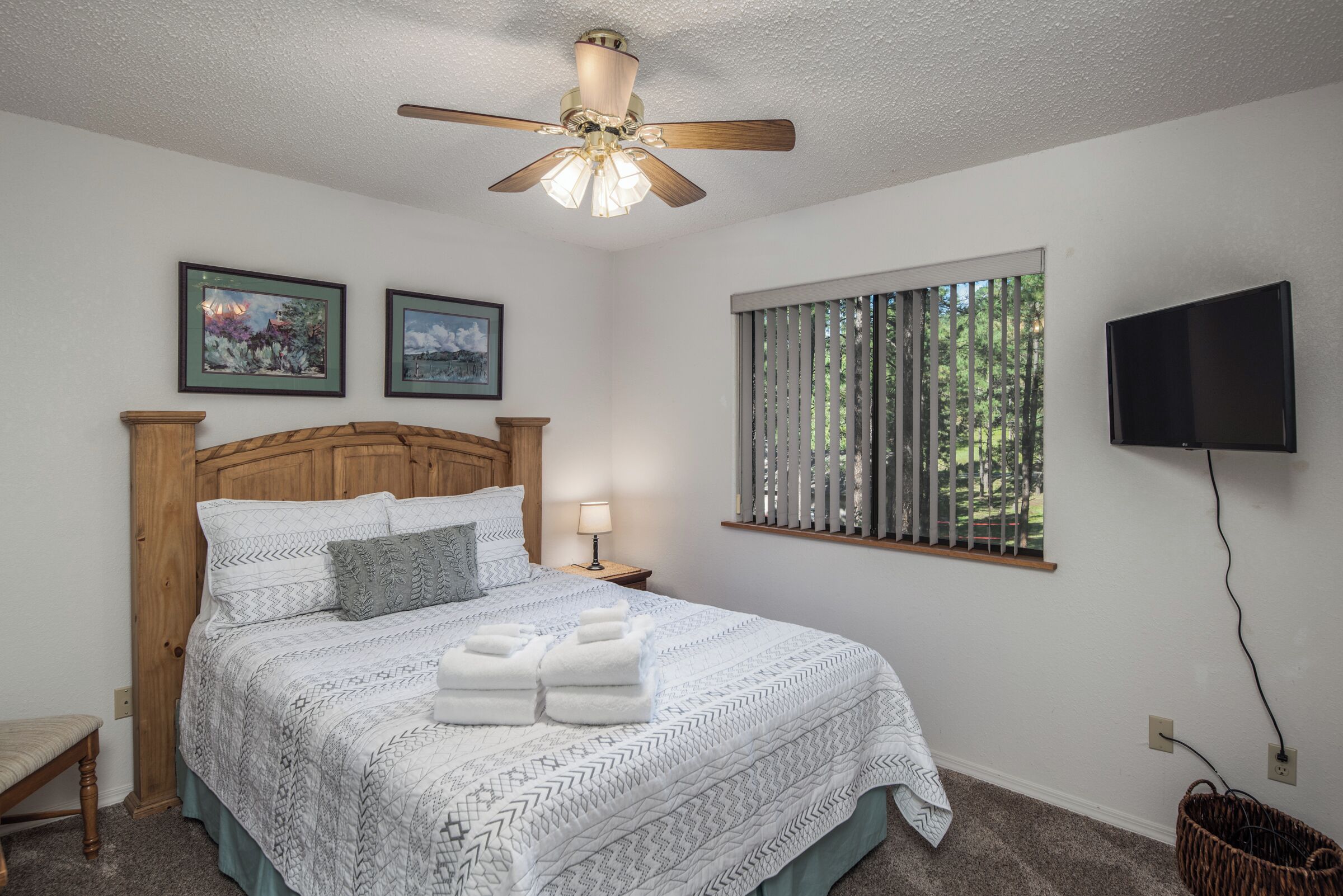 A bedroom with a wooden headboard, a bed with white linens and a patterned blanket, a ceiling fan, a window with blinds, and a mounted TV.