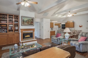 A spacious living area with a fireplace, a dining table, and comfortable seating. There is a kitchen in the background with wooden cabinets and a ceiling fan.