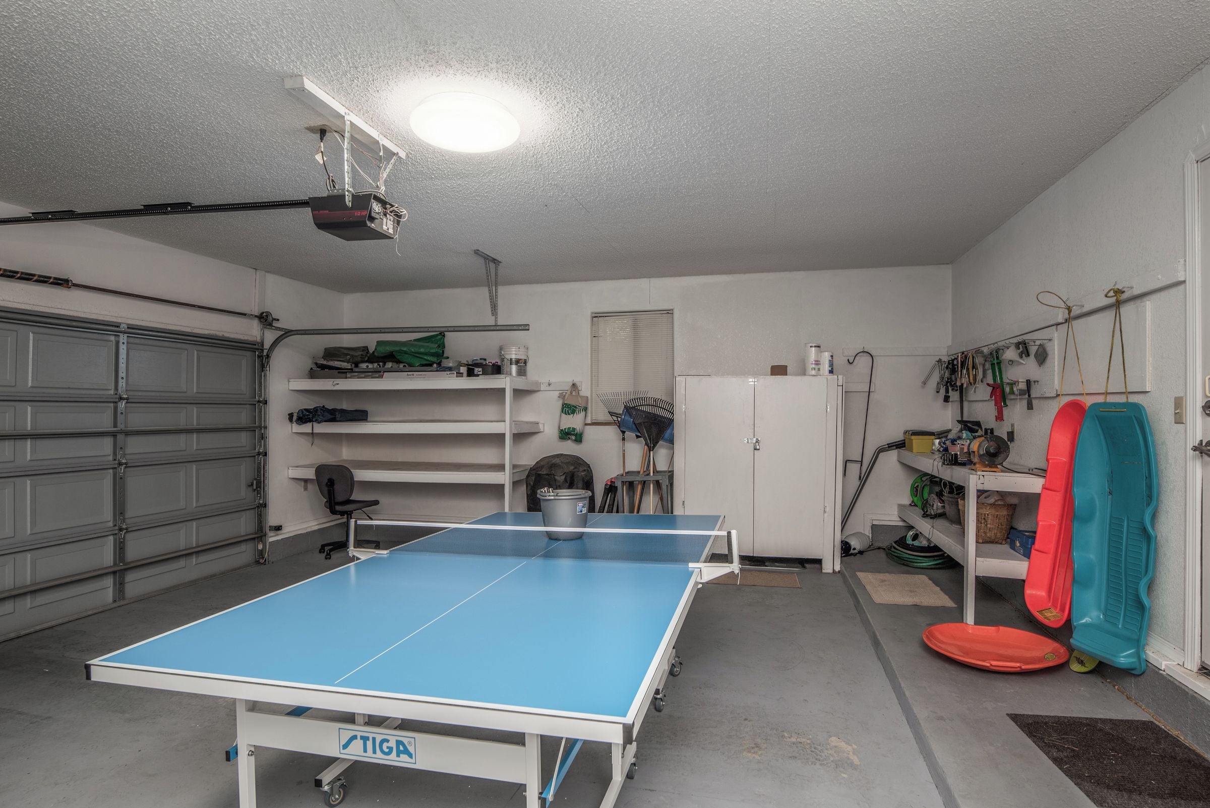 A garage with a ping pong table, shelves with tools, a refrigerator, and a door with a towel hanging.