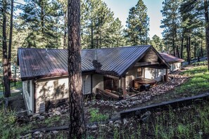 A cabin with a metal roof, wooden deck, and a stone foundation, surrounded by trees.