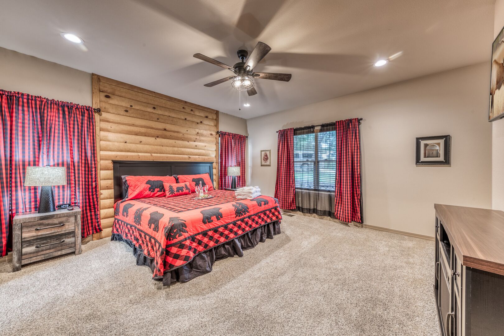 A bedroom with a wooden log wall, a bed with a red and black checkered bedspread, a ceiling fan, and a window with red curtains.