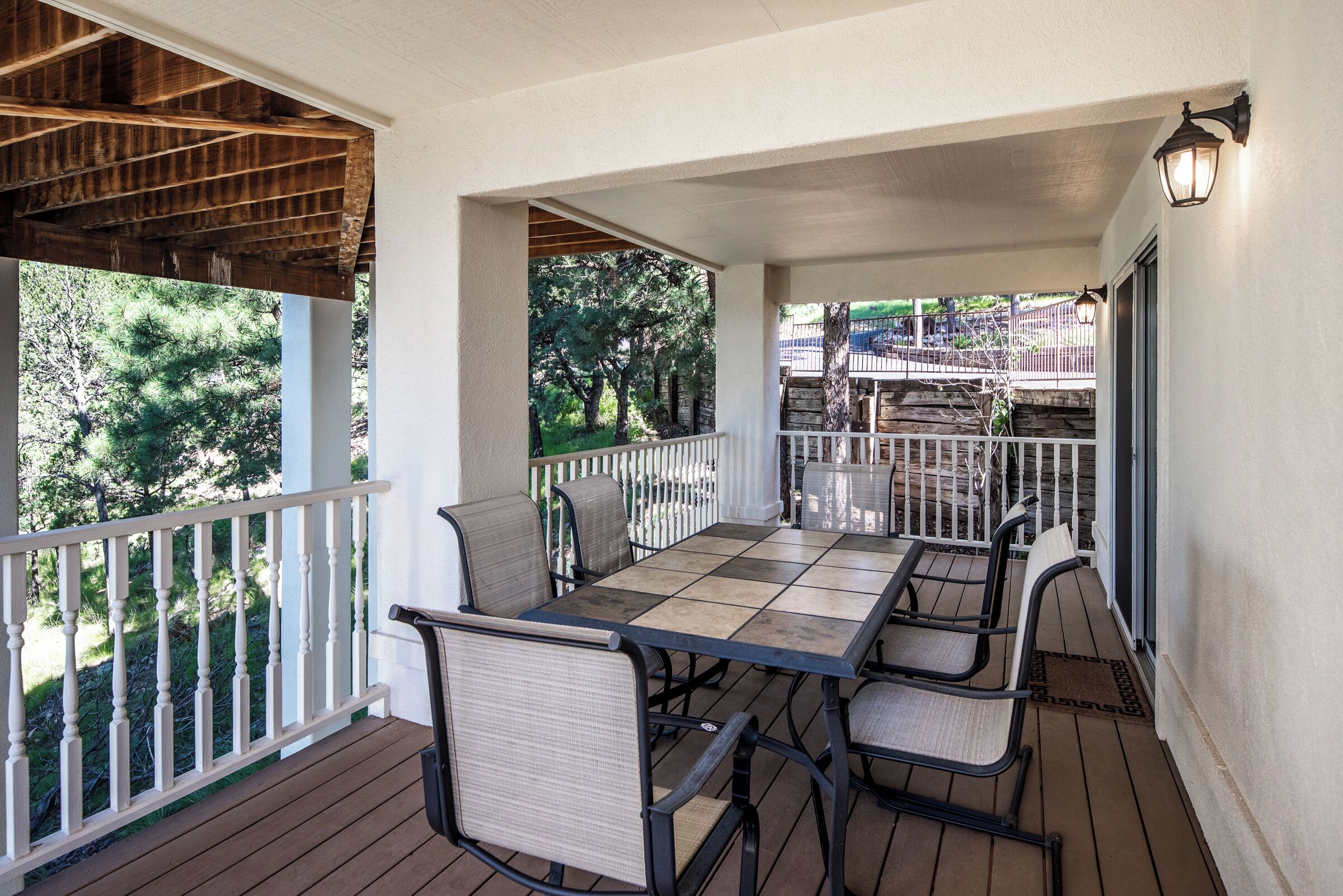 A covered outdoor patio with a wooden table and chairs, white railings, and a view of trees and a playground.