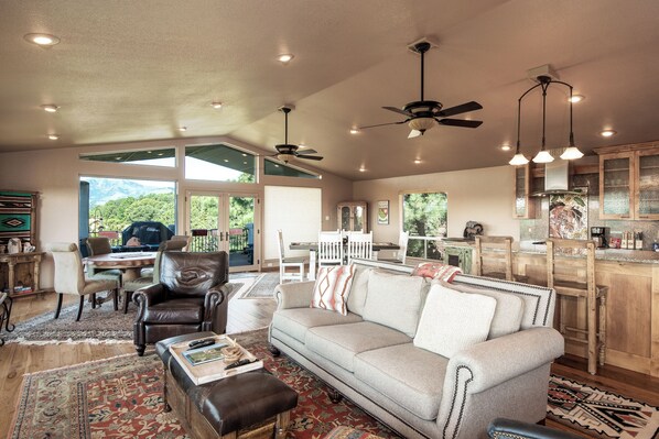 A spacious living area with a sofa, chairs, a coffee table, and a dining table. There are ceiling fans, pendant lights, and a view of the outdoors. - Alto Grand View by Ruidoso Vacation Rentals (Alto)