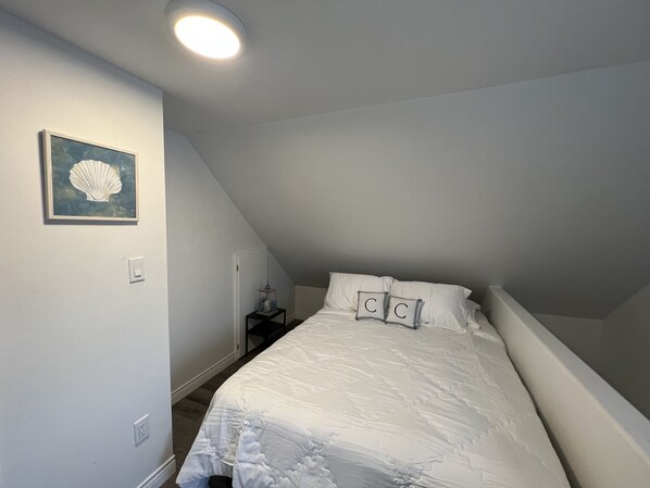 A bedroom with a sloped ceiling, a bed with white bedding, a framed picture on the wall, and a small bedside table with a lamp. - Cricker's Corner Vacation Home - North Rustico (North Rustico)