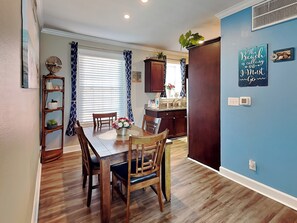 A dining area with a wooden table and chairs, a kitchen with dark cabinets, and a blue accent wall with a beach-themed sign. - Dawn 223 - Buena Vista by Ryson Vacation Rentals (Galveston)