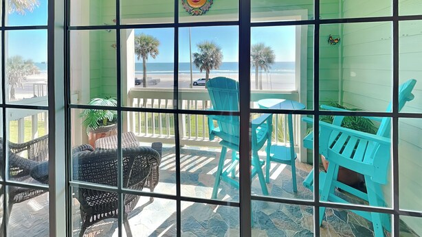 Een balkon met turquoise stoelen en een rieten stoel, met uitzicht op een strand met palmbomen en een heldere lucht.