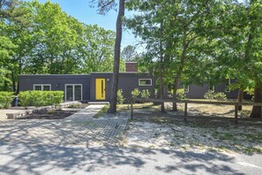 Une maison moderne avec une allée pavée, une porte jaune et une cour clôturée avec des arbres.