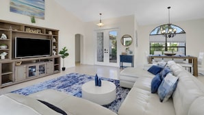 Un salon spacieux avec un canapé blanc, une table basse et un téléviseur posé sur un meuble en bois. Un tapis à motifs bleus et blancs recouvre le sol.