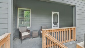 Une véranda couverte avec une balustrade en bois, deux fauteuils en osier, une petite table et une fenêtre avec des stores.