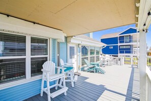 A covered deck with white Adirondack chairs and a small table, overlooking a blue building with a balcony.
