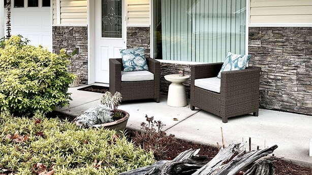 A covered porch with wicker furniture, a stone accent wall, and a potted plant. - Modern, cozy home in private beach community Admirals Cove (Coupeville)