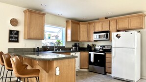 A modern kitchen with wooden cabinets, a central island with bar stools, a white refrigerator, and a built-in oven. - Modern, cozy home in private beach community Admirals Cove (Coupeville)
