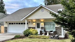 A single-story house with a garage, a covered porch with outdoor seating, and a well-maintained lawn. - Modern, cozy home in private beach community Admirals Cove (Coupeville)