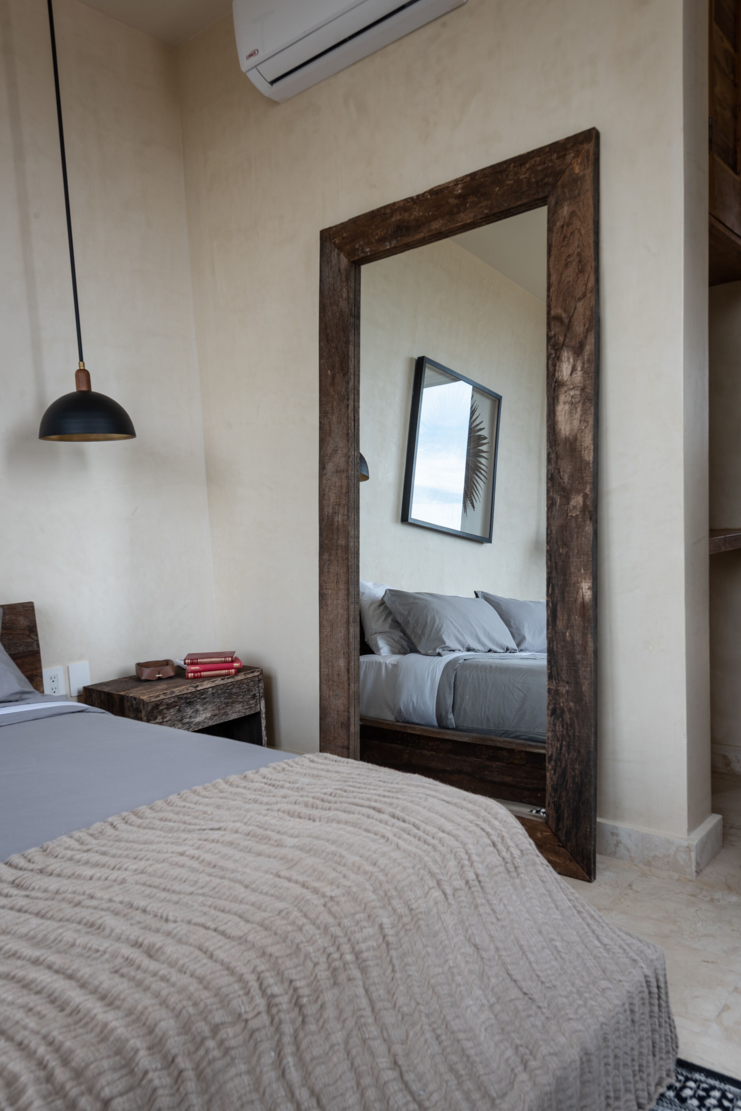 A bedroom with a large mirror, a bed with a grey blanket, a bedside table with books, and a pendant light.