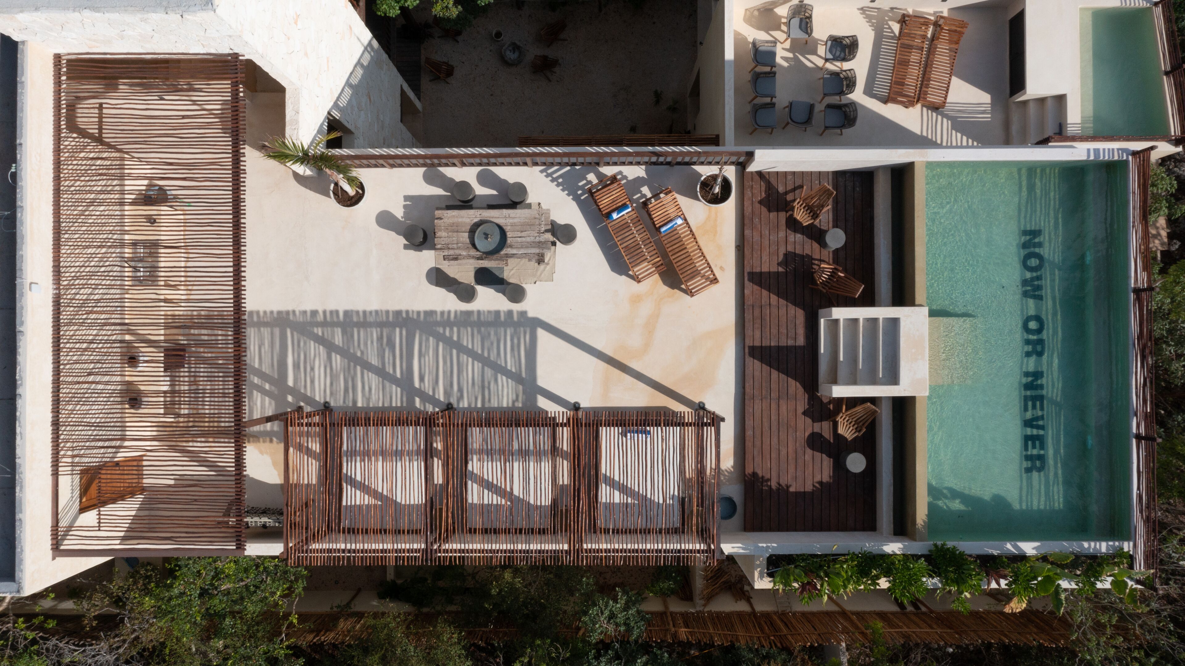 An aerial view of a rooftop pool area with lounge chairs, a wooden deck, and a sign that reads "NOW OR NEVER".