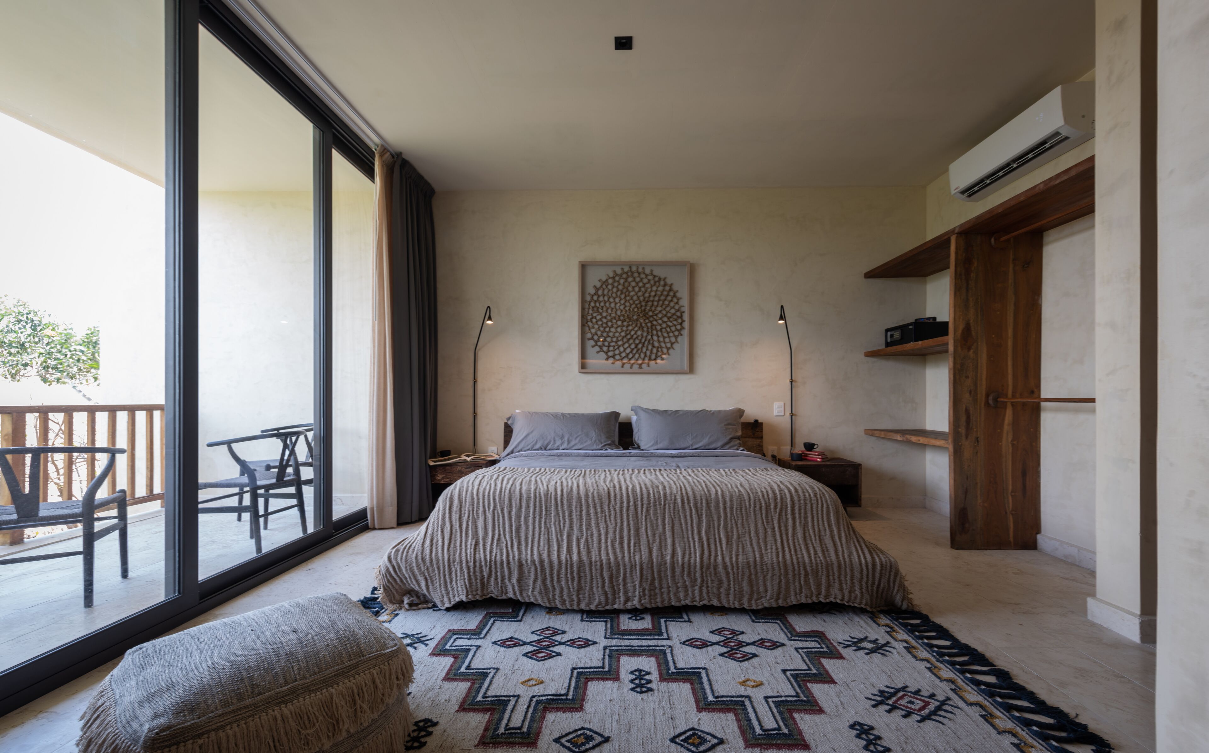 Une chambre avec une grande fenêtre, un lit recouvert d’une couvre-lit à motifs, un tapis aux motifs géométriques et un banc en bois.