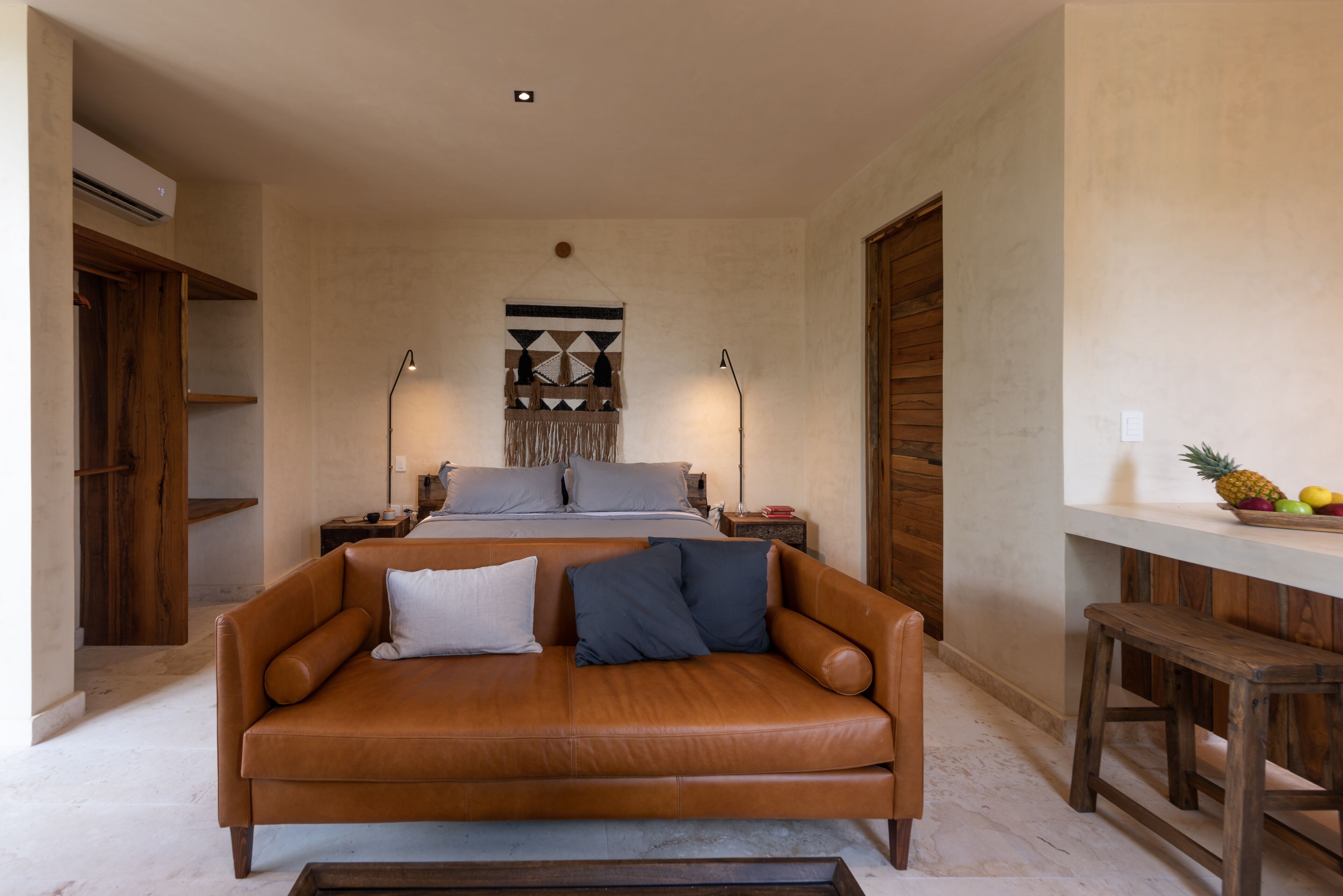 A bedroom with a bed, a wooden wardrobe, a nightstand, a lamp, and a small table with fruits.