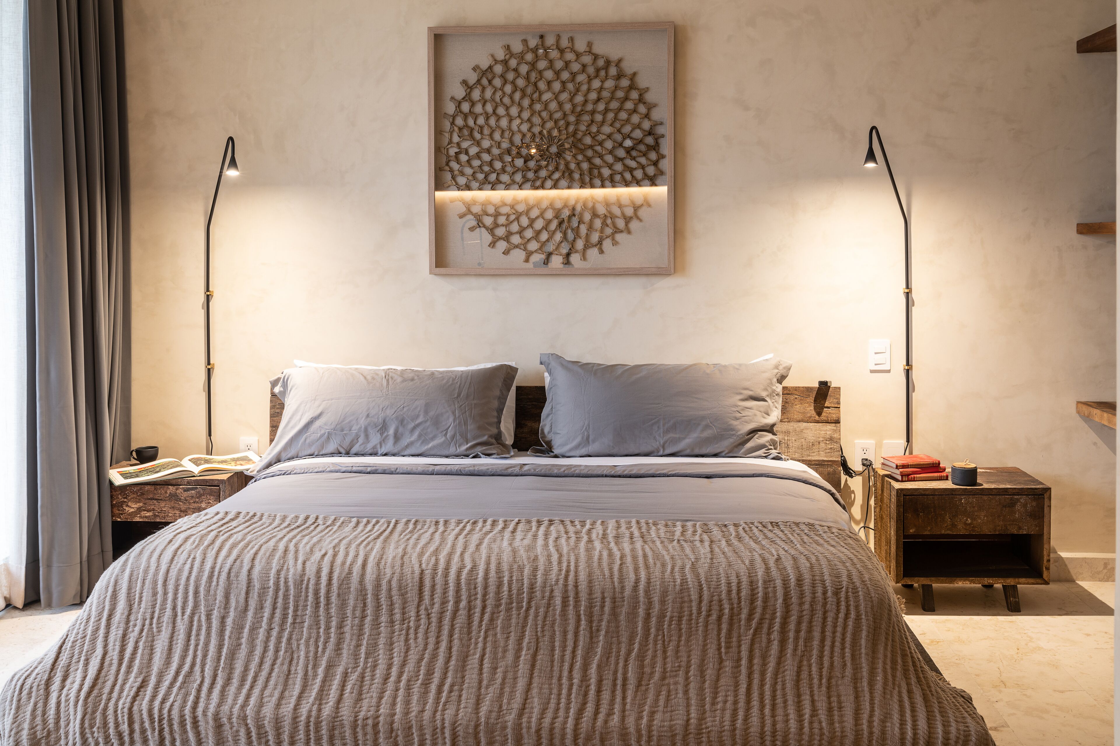 Un lit bien fait, recouvert d’une couverture à texture intéressante, deux lampes de chevet, une œuvre d’art abstraite accrochée au mur, et une petite table de chevet en bois sur laquelle reposent un livre et une tasse.