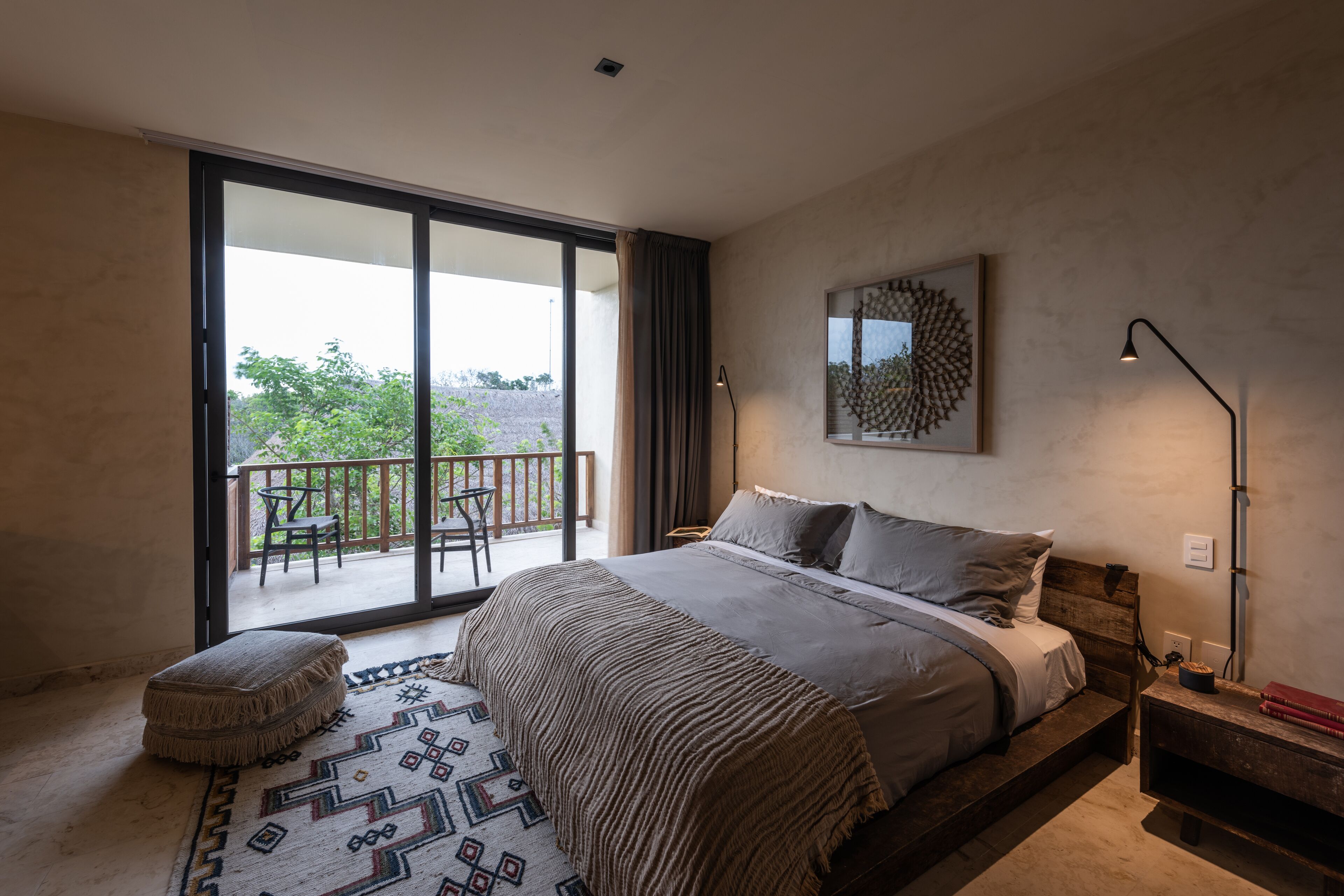 A bedroom with a large bed, a balcony, a patterned rug, and a wall-mounted light fixture.