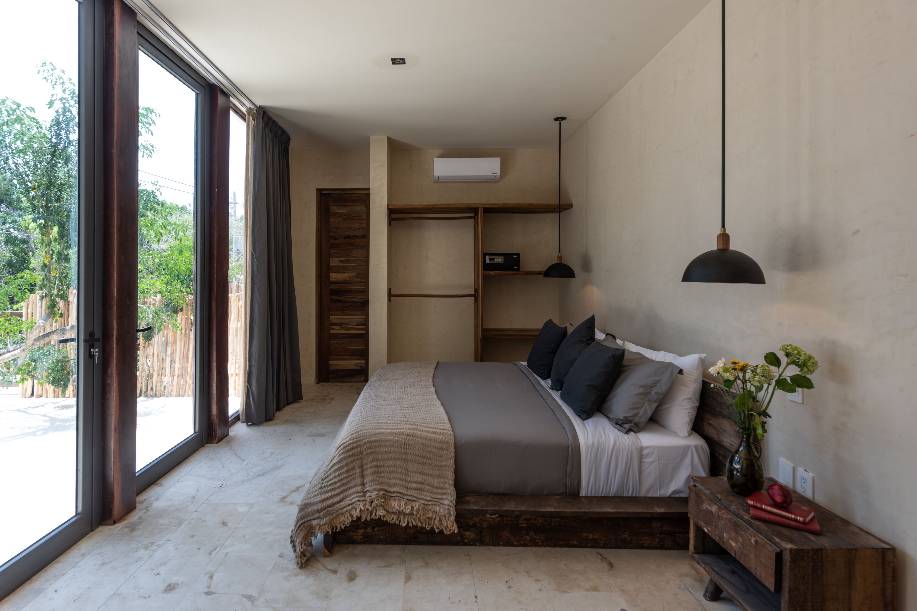 A bedroom with a large window, a bed with a grey blanket, a bedside table with a vase, and a wooden bench.
