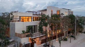 A modern multi-story building with a mix of balconies and terraces, surrounded by trees and a sandy ground. - Unho Tulum (Tulum)