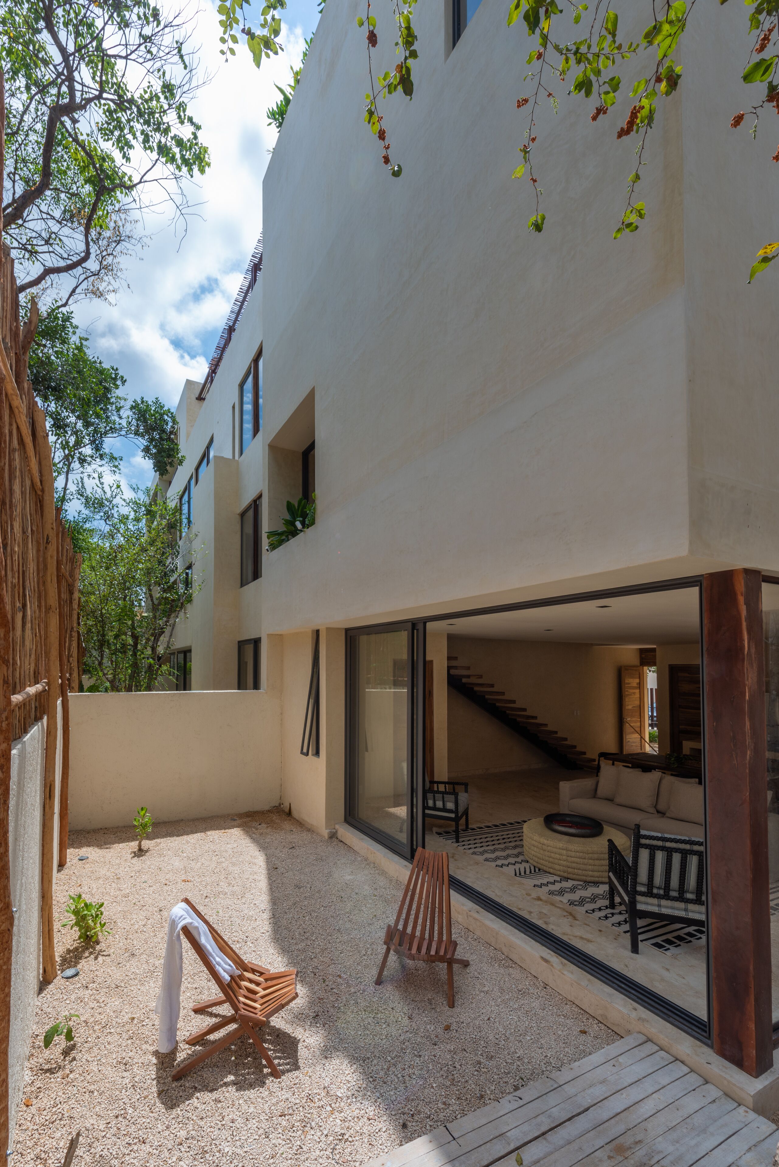 Un edificio moderno con un espacio exterior cubierto que cuenta con una terraza de madera, una zona de estar con un sofá y una mesa de centro, y una escalera que conduce a un nivel superior.