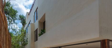 Un edificio moderno con un espacio exterior cubierto que cuenta con una terraza de madera, una zona de estar con un sofá y una mesa de centro, y una escalera que conduce a un nivel superior.