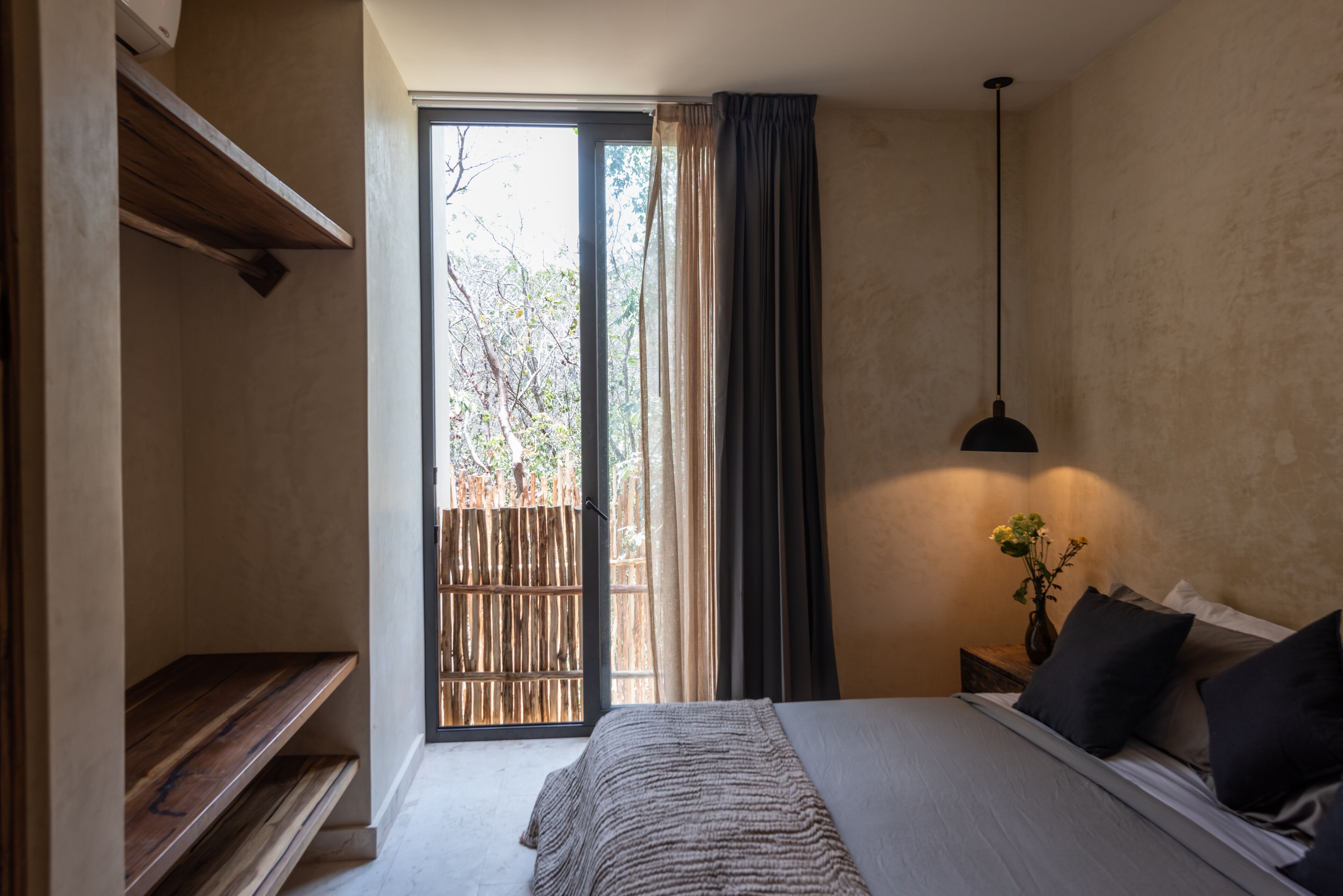 Quarto com cama, mesa de cabeceira, vaso com flores, luminária pendente e vista para árvores pela janela.