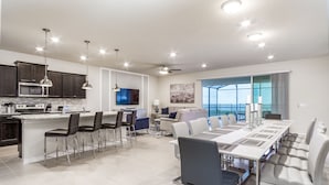A modern kitchen with a central island, bar stools, a dining area with a long table and chairs, and a large window offering a view.