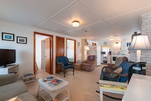 Relax in this open-plan living area with a sofa, chair, and bench, featuring a TV, coffee table, and kitchenette with a refrigerator, stove, and sink.