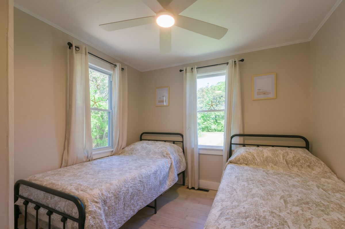 Une chambre avec deux lits simples, un ventilateur de plafond et des fenêtres équipées de rideaux.