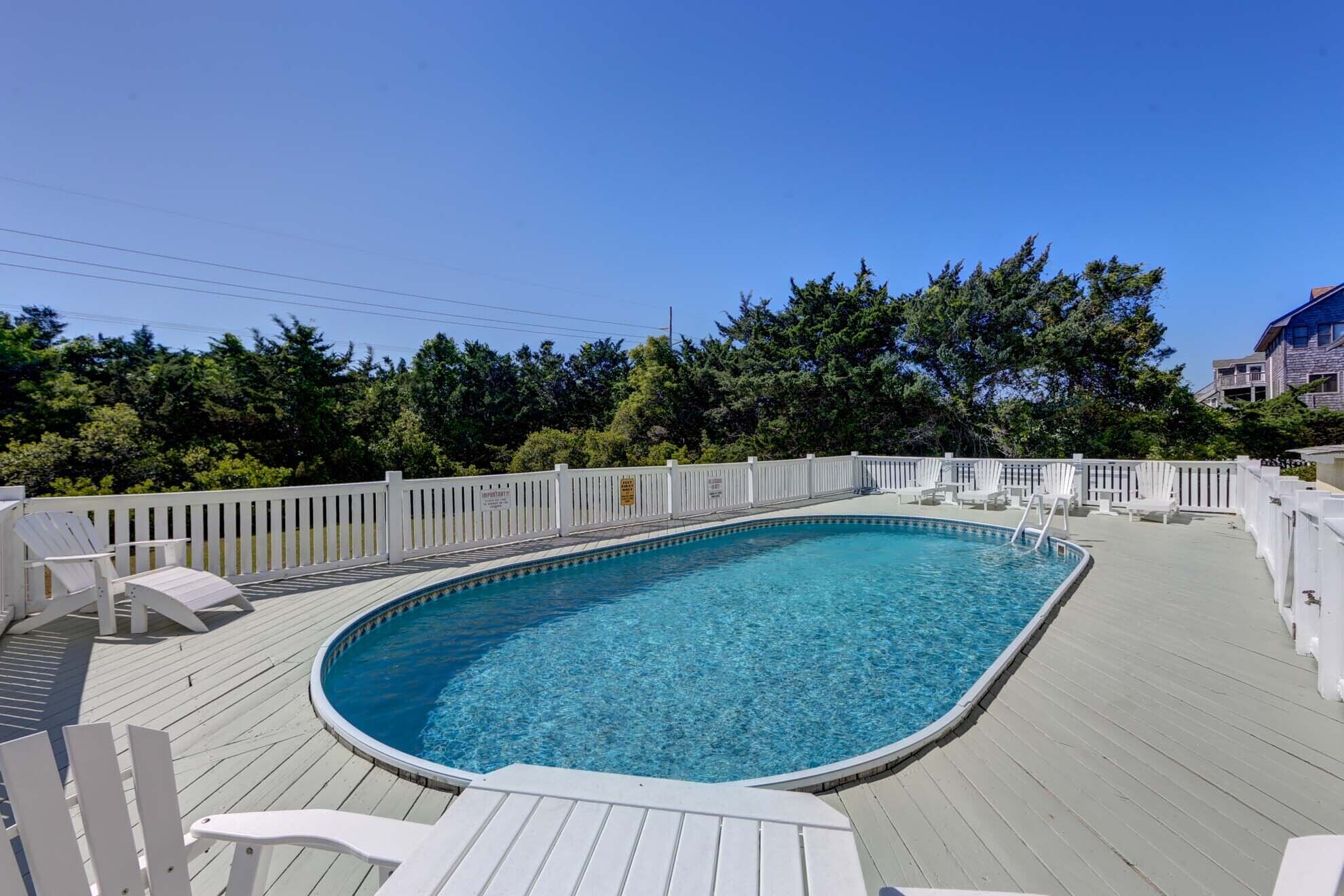 Una piscina exterior con muelle de madera, rodeada de vallas blancas y árboles.