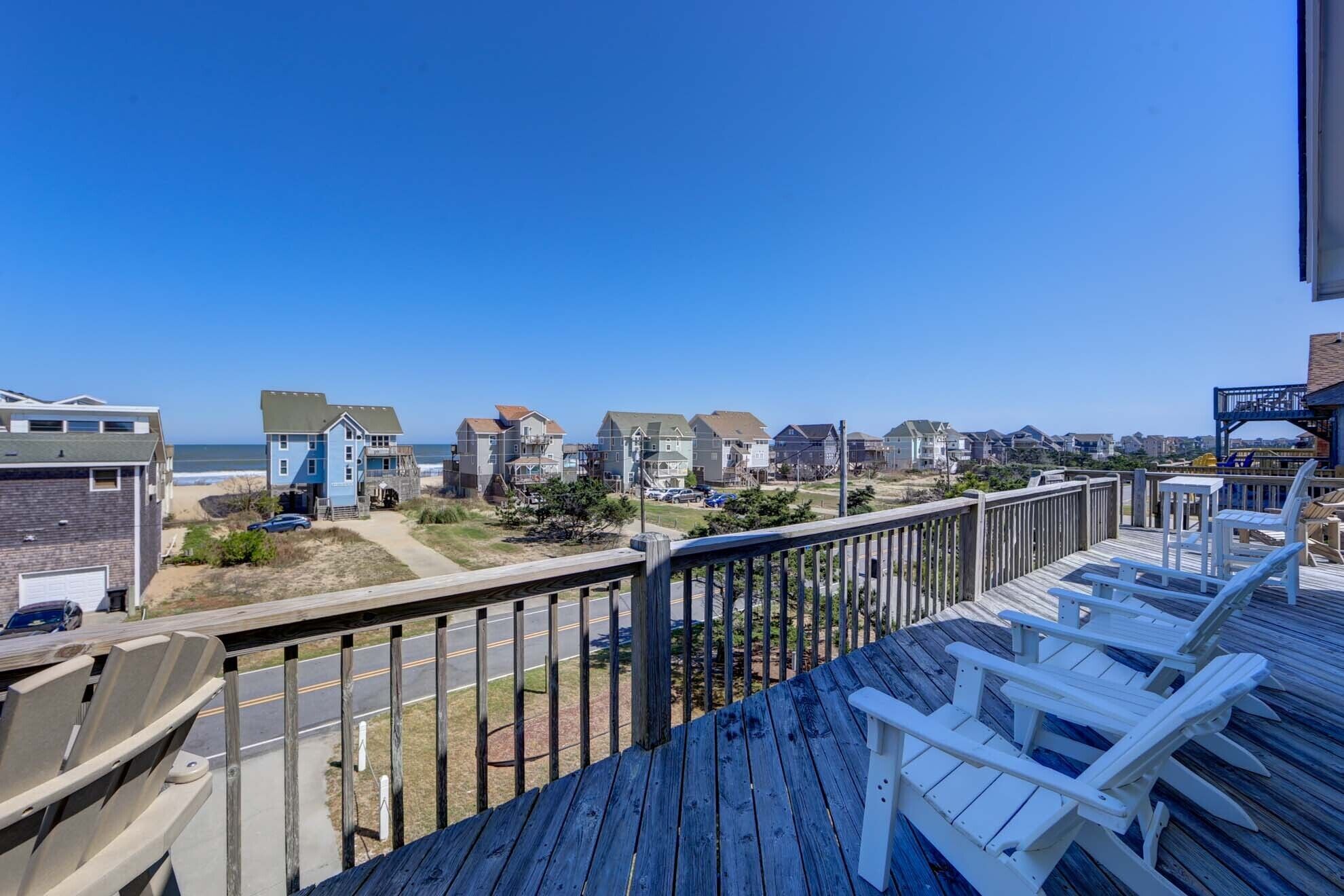 A wooden deck with white chairs overlooking a beach and residential area.