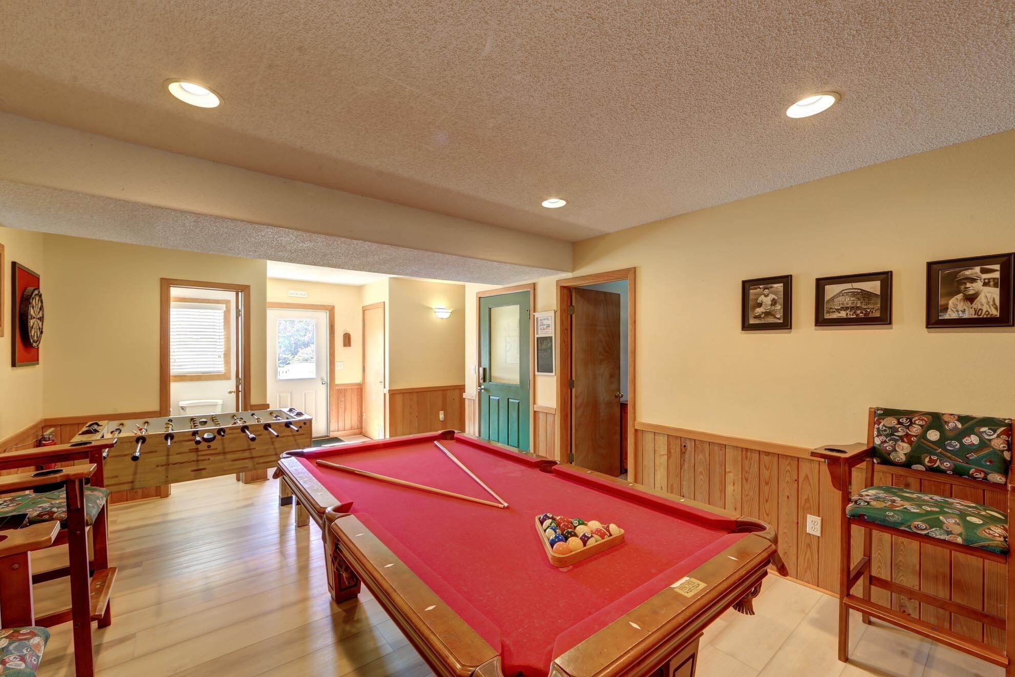 A pool table with a red felt surface, a foosball table, and a billiards table in a room with wooden paneling and recessed ceiling lights.