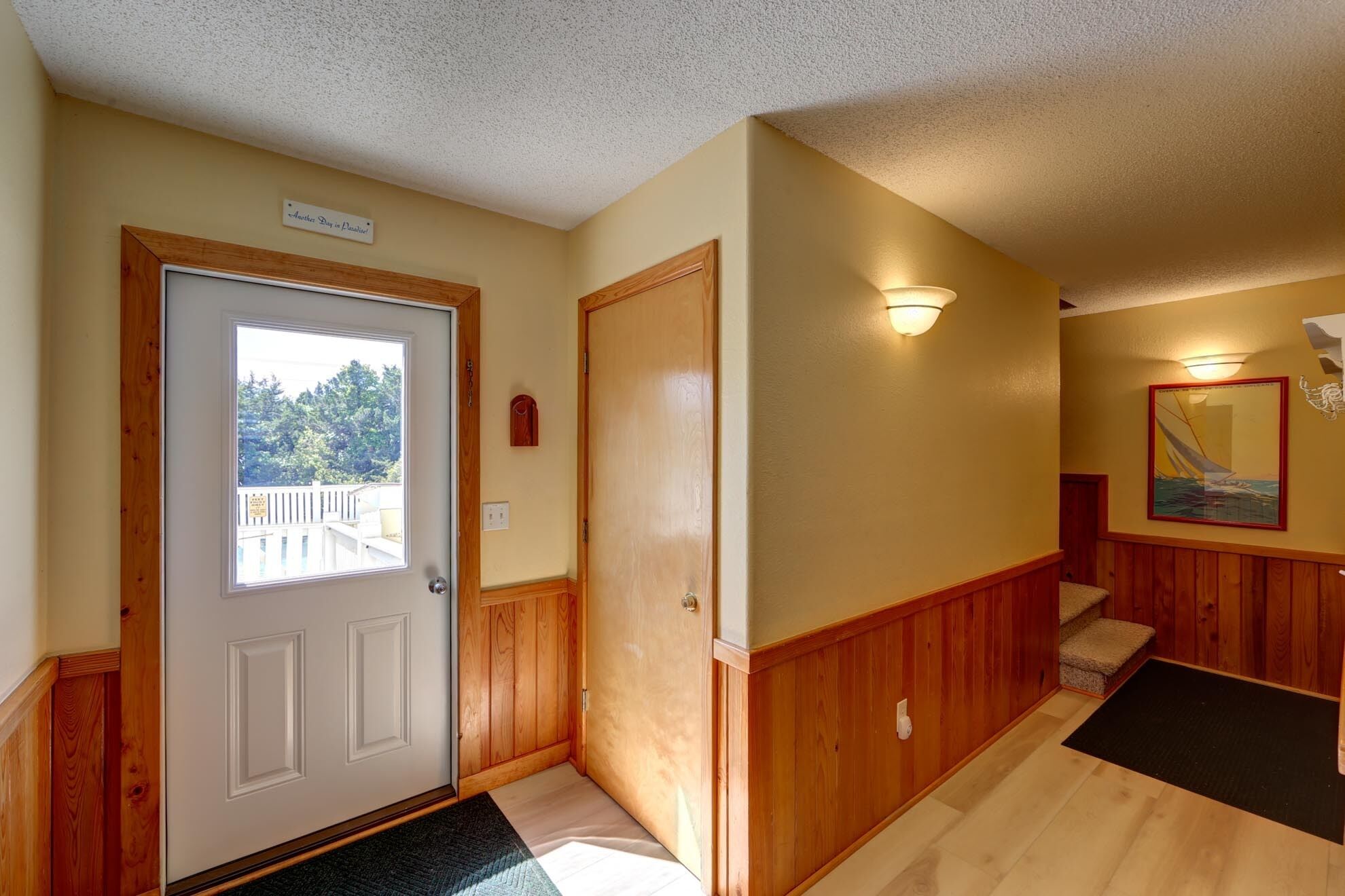 A hallway with a wooden door, a small wooden bench, a painting, and a light fixture.