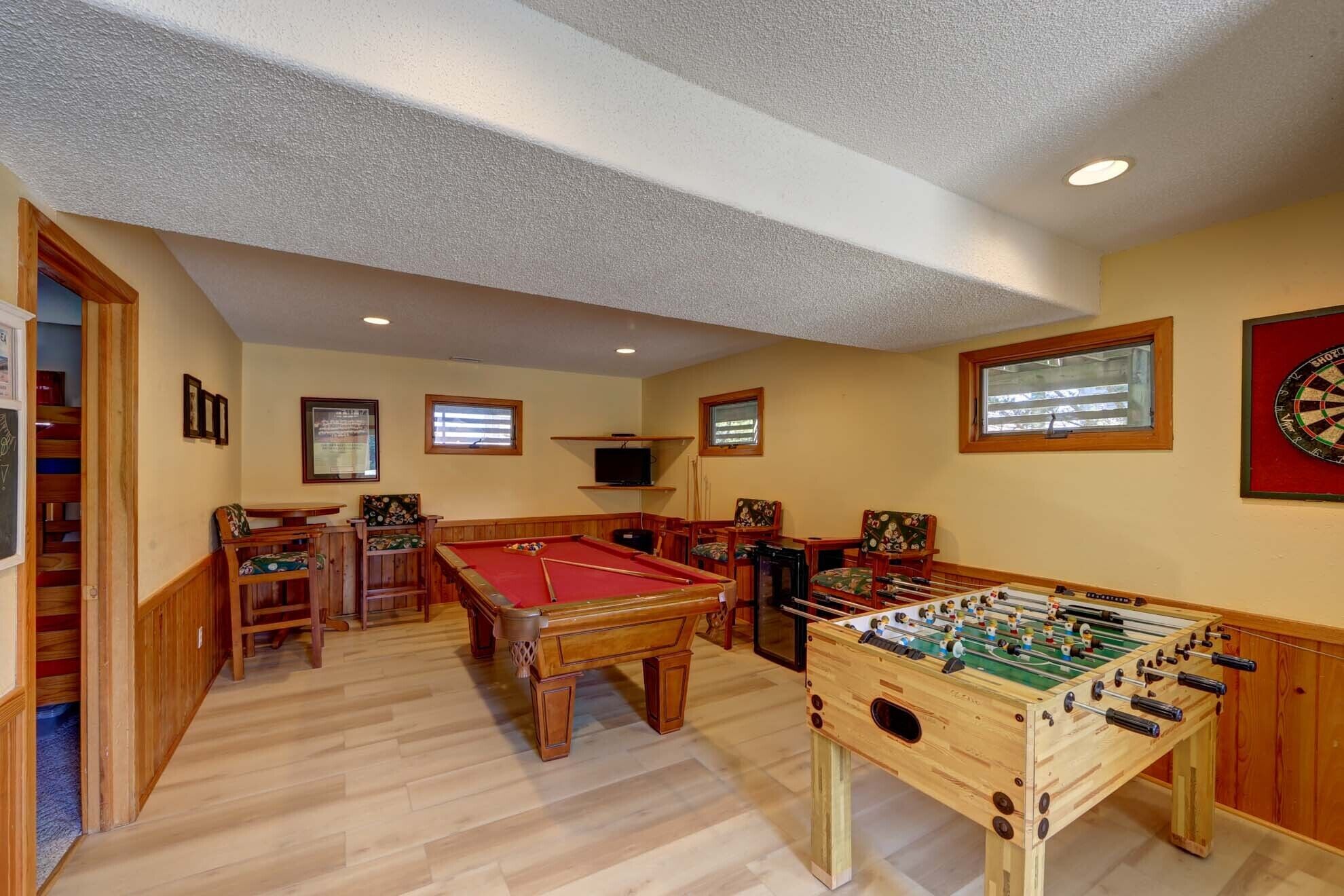 A room with a pool table, foosball table, and dartboard. There are wooden chairs and a television mounted on the wall.