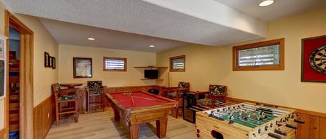 A room with a pool table, foosball table, and dartboard. There are wooden chairs and a television mounted on the wall.