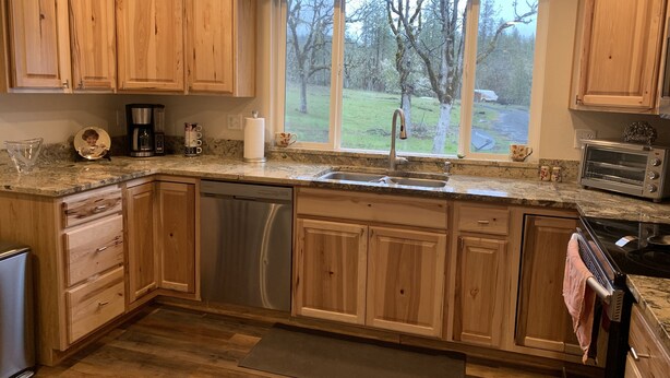 A kitchen with wooden cabinets, a dishwasher, a sink, a window with a view of trees, and a toaster. - Spectacular Cozy Cottage- Wood Stove, Fire Pit, Creek (Shady Cove)