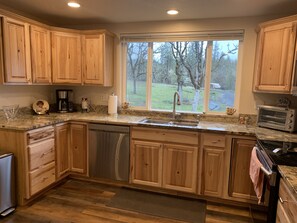 A kitchen with wooden cabinets, a dishwasher, a sink, a window with a view of trees, and a toaster. - Spectacular Cozy Cottage- Wood Stove, Fire Pit, Creek (Shady Cove)