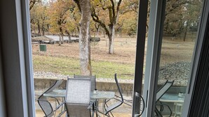 A view through a glass door to an outdoor patio with a table and chairs, a doormat, and a grassy area with trees. - Spectacular Cozy Cottage- Wood Stove, Fire Pit, Creek (Shady Cove)