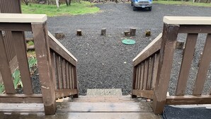 A wooden deck leading to a gravel driveway with a blue truck parked in the distance. - Spectacular Cozy Cottage- Wood Stove, Fire Pit, Creek (Shady Cove)